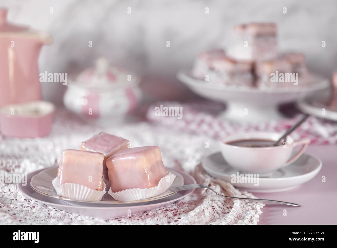 Kleine rosa glasierte Kuchen auf einem Teller neben einer Kaffeetasse und feinem Porzellangeschirr Stockfoto