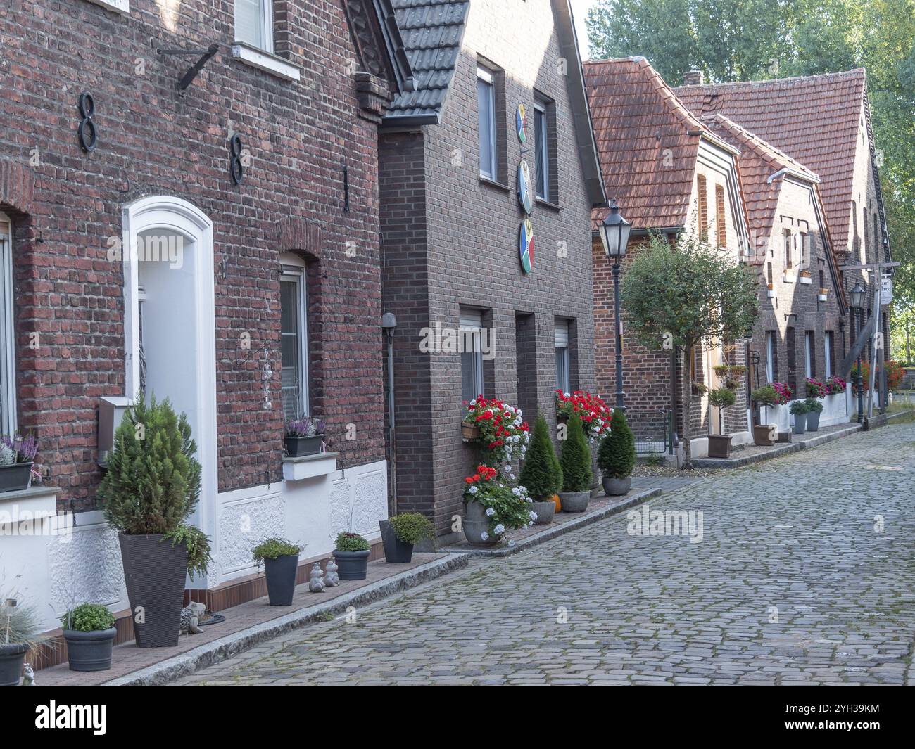 Reihe von Backsteinhäusern mit dekorativen Blumen und Pflanzen in Töpfen entlang einer Kopfsteinpflasterstraße an einem sonnigen Tag, Krudenburg, Hünxe, Nordrhein-Westfalen, Keime Stockfoto