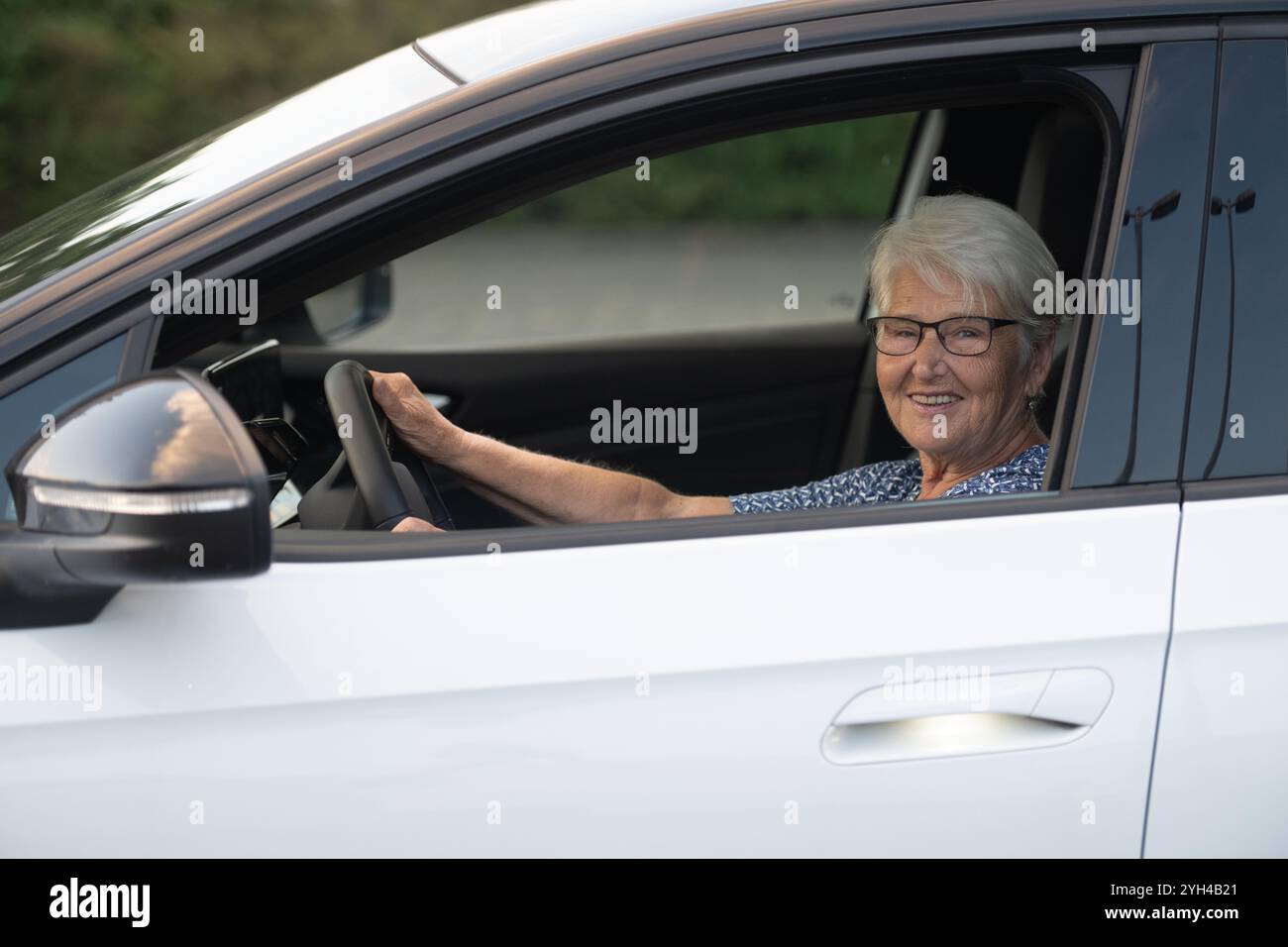 Eine fröhliche ältere Frau mit Brille sitzt hinter dem Steuer eines weißen Autos und hält selbstbewusst das Lenkrad, während sie ihre Fahrt genießt. Stockfoto