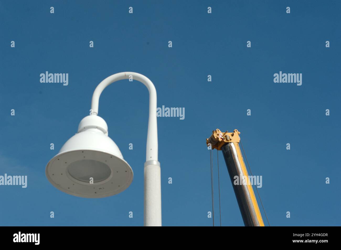 Isolierter weißer, gekrümmter Lampenpfosten mit Haken und Spitze eines gelben Krans vor einem hellblauen Himmel. Keine Leute und Platz zum Kopieren. Stockfoto