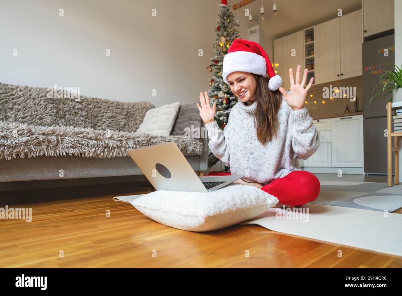 Junge Frau mit Weihnachtsmann-Hut in Lotuspose, Videoanruf Freunde auf Laptop, lächelnd und winkend. Stockfoto