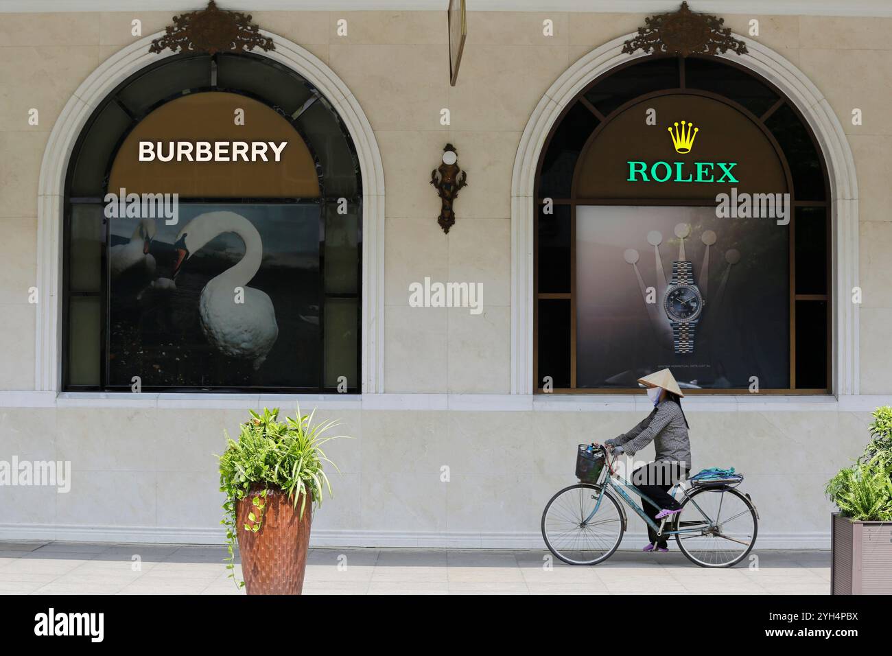 Hanoi, Vietnam - 10. Juli 2023: Eine Vietnamesin, die den traditionellen Non-La-Hut trägt, fährt mit dem Fahrrad vorbei an Burberry- und Rolex-Geschäften. Stockfoto