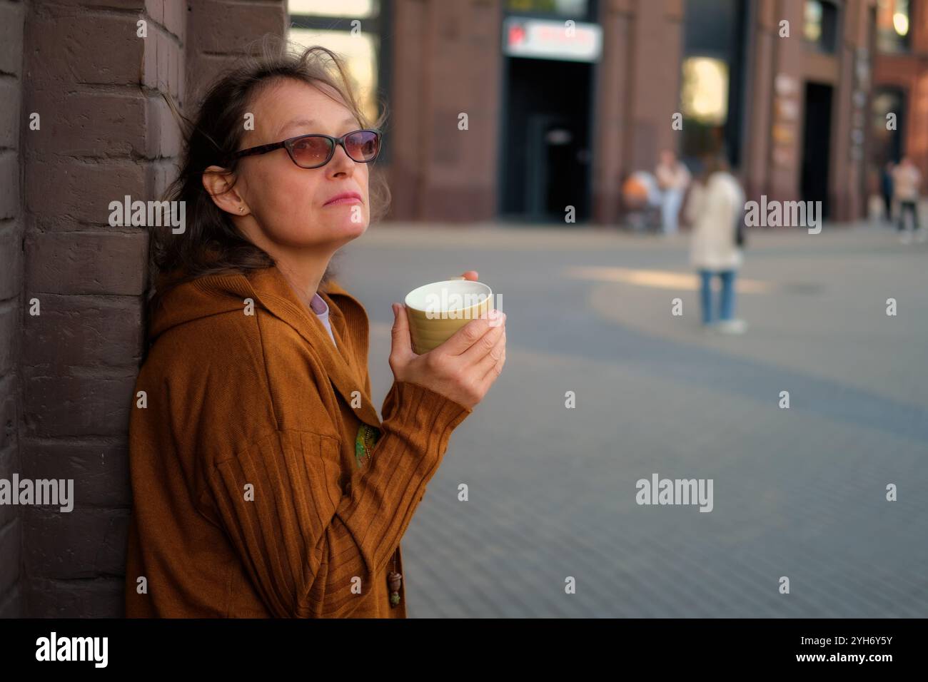 Eine Frau mittleren Alters in Sonnenbrille, die einen Becher hält und sich an eine Ziegelmauer auf einer Stadtstraße lehnt. Urbanes Lifestyle-Konzept. Frau 50 Jahre alt (50 Y Stockfoto