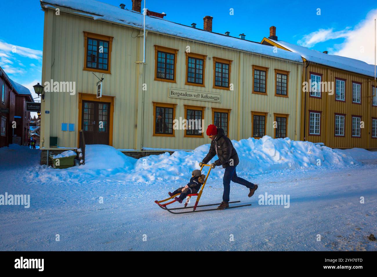 Ein Vater fährt mit seiner Tochter auf einem Schneeschlitten durch die verschneiten Straßen von Røros an einem klaren, sonnigen Tag und genießt den Charme dieses Winterwunderlandes. Stockfoto