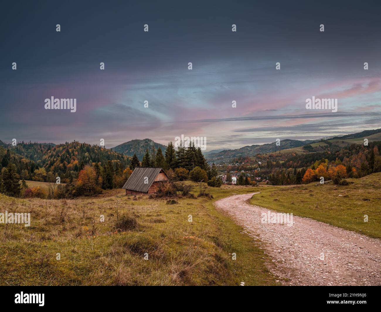 Idyllische Herbstlandschaft mit einer rustikalen Hütte inmitten von sanften Hügeln, lebhaftem Laub und einem gewundenen Pfad unter einem dramatischen, farbenfrohen Himmel. Perfekt für Stockfoto