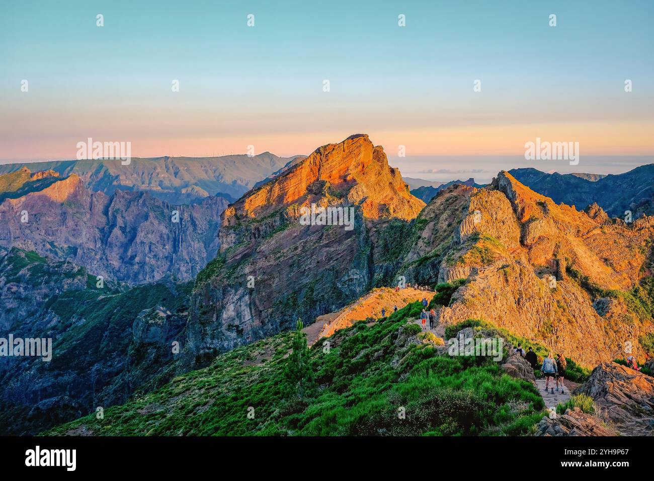 Ein Blick auf den Sonnenaufgang von einem Berggipfel mit Wanderern auf einem malerischen Pfad, umgeben von zerklüftetem felsigem Gelände. Stockfoto