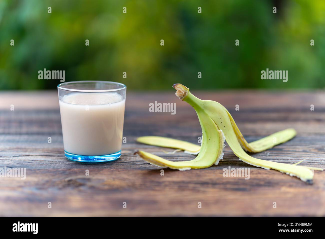 Bananensaft in einer blauen Glasschale und die Bananenschale auf einem Holztisch auf einem grünen, verschwommenen Hintergrund der Natur Stockfoto
