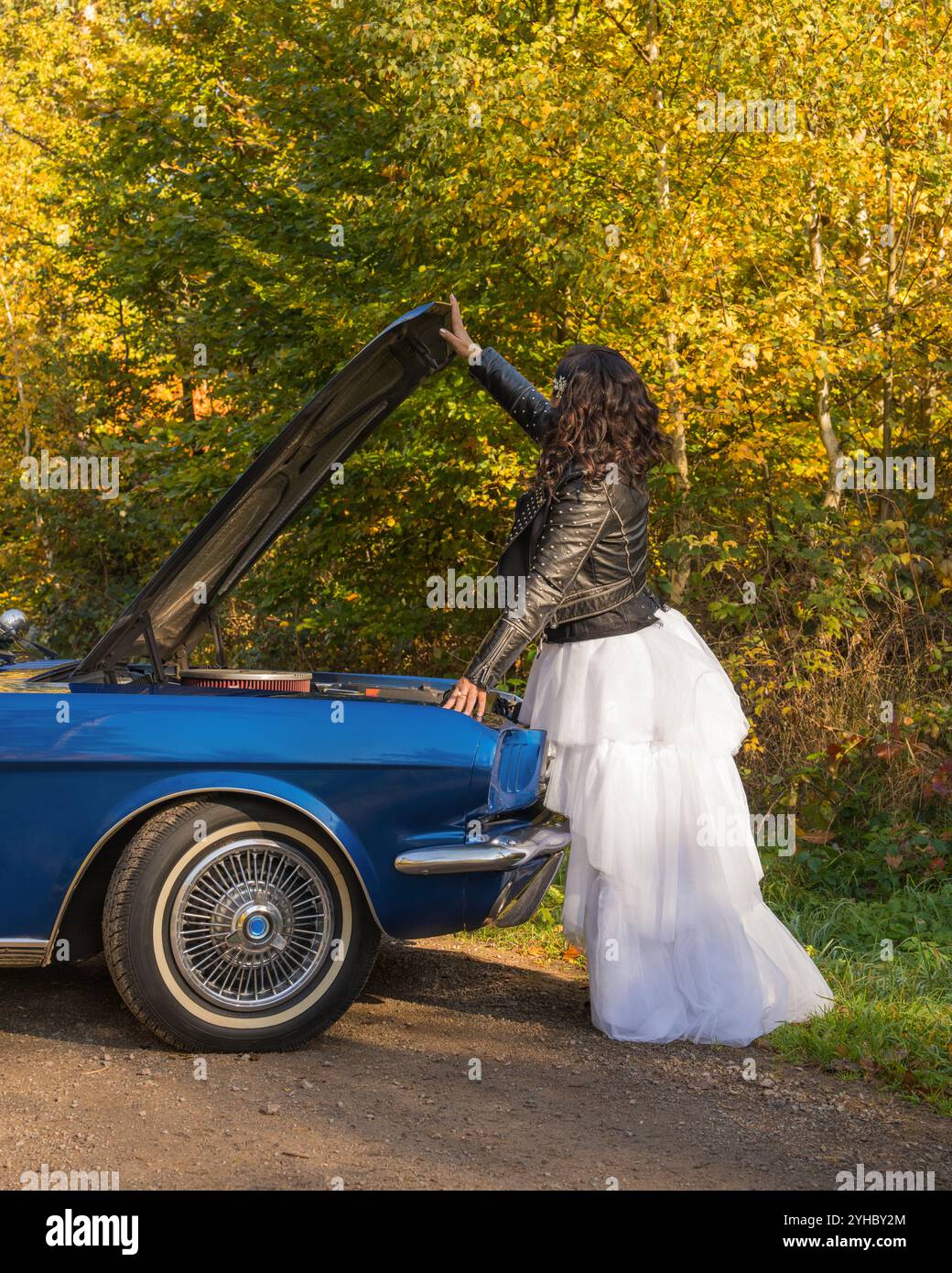 Schönes Mädchen in Lederjacke, weißem geschwollenen Rock, Stiefeln in der Nähe des Retro-Autos im Herbstwald. Frau in der Nähe eines Oldtimers mit offener Motorhaube. Stockfoto