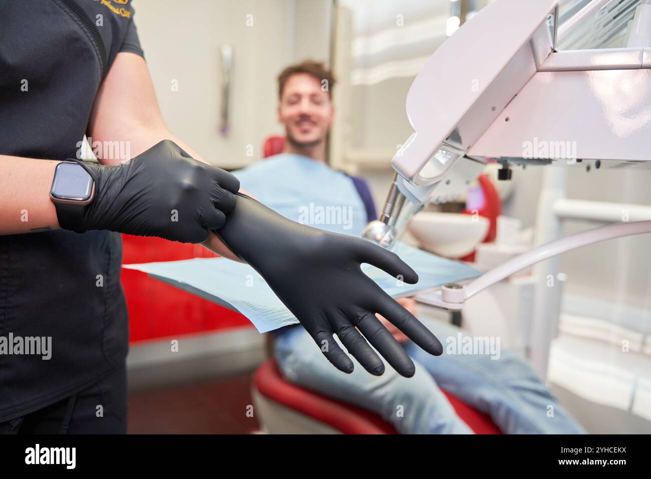 Junger lateinischer Zahnarzt in blau gekleidet, Details an Handschuhen, Hintergrund der Zahnarztkabine mit sitzendem Klienten, nicht erkennbares Modell Stockfoto