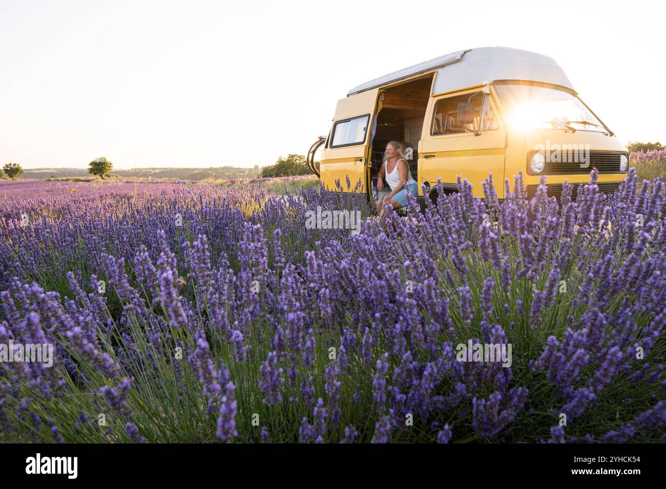 Junge blonde Frau genießt die Aussicht in ihrem gelben Retro-Wohnwagen bei Sonnenaufgang in Valensole Lavendelfeldern in der Provence, Südfrankreich Stockfoto