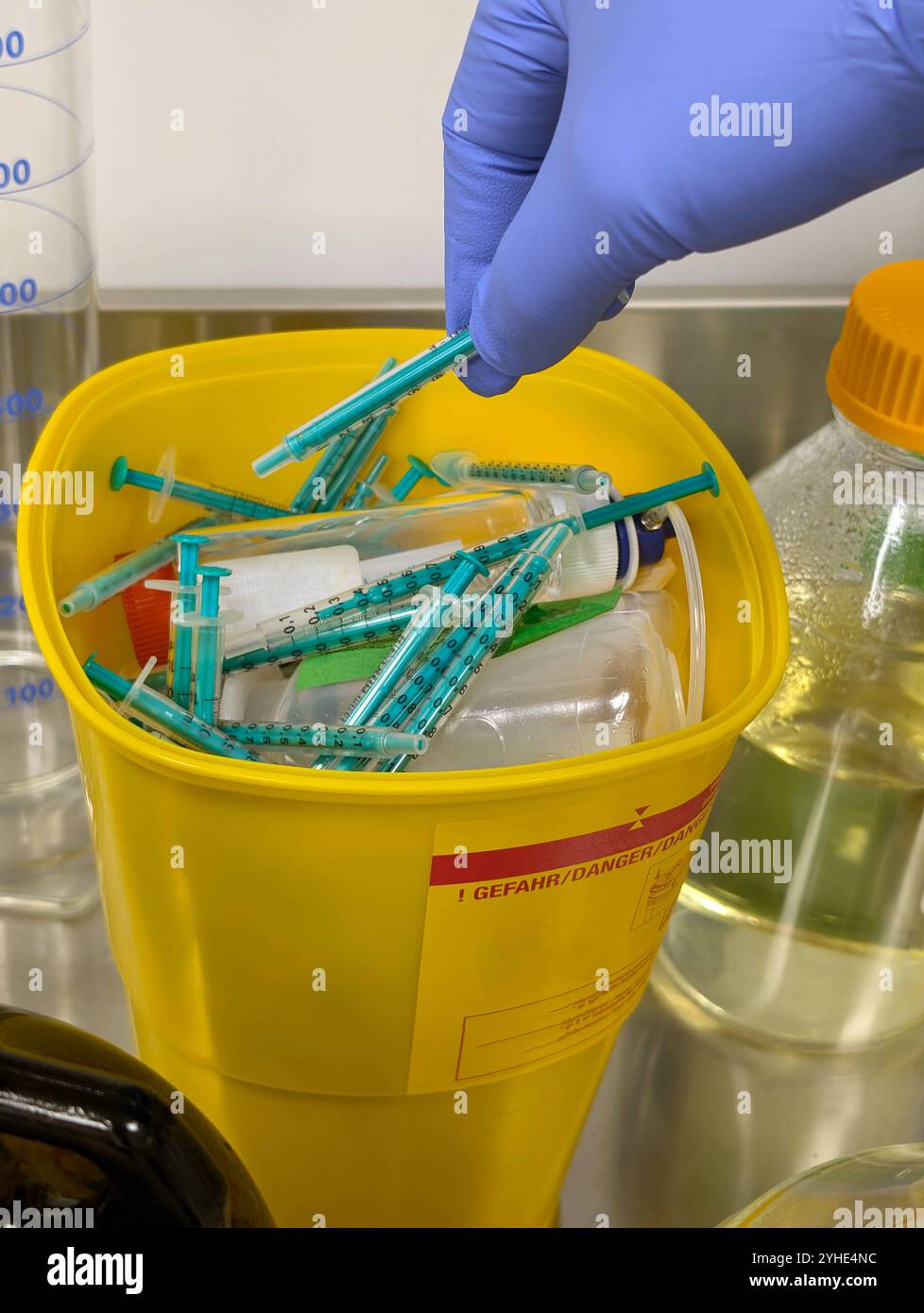 Eine gelbe Abfallbox mit der Aufschrift Gefahr, gefüllt mit gebrauchten Spritzen in einem Labor und einer Hand mit einem blauen Schutzhandschuh, die eine weitere Spritze in die Schachtel legt Stockfoto