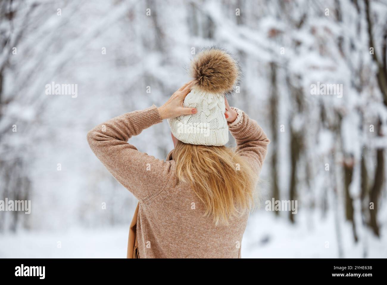 Eine Frau mit Pullover zieht während der Wanderung im verschneiten Wald einen gestrickten Bommelmütze an. Warme Kleidung bei kaltem Winterwetter Stockfoto