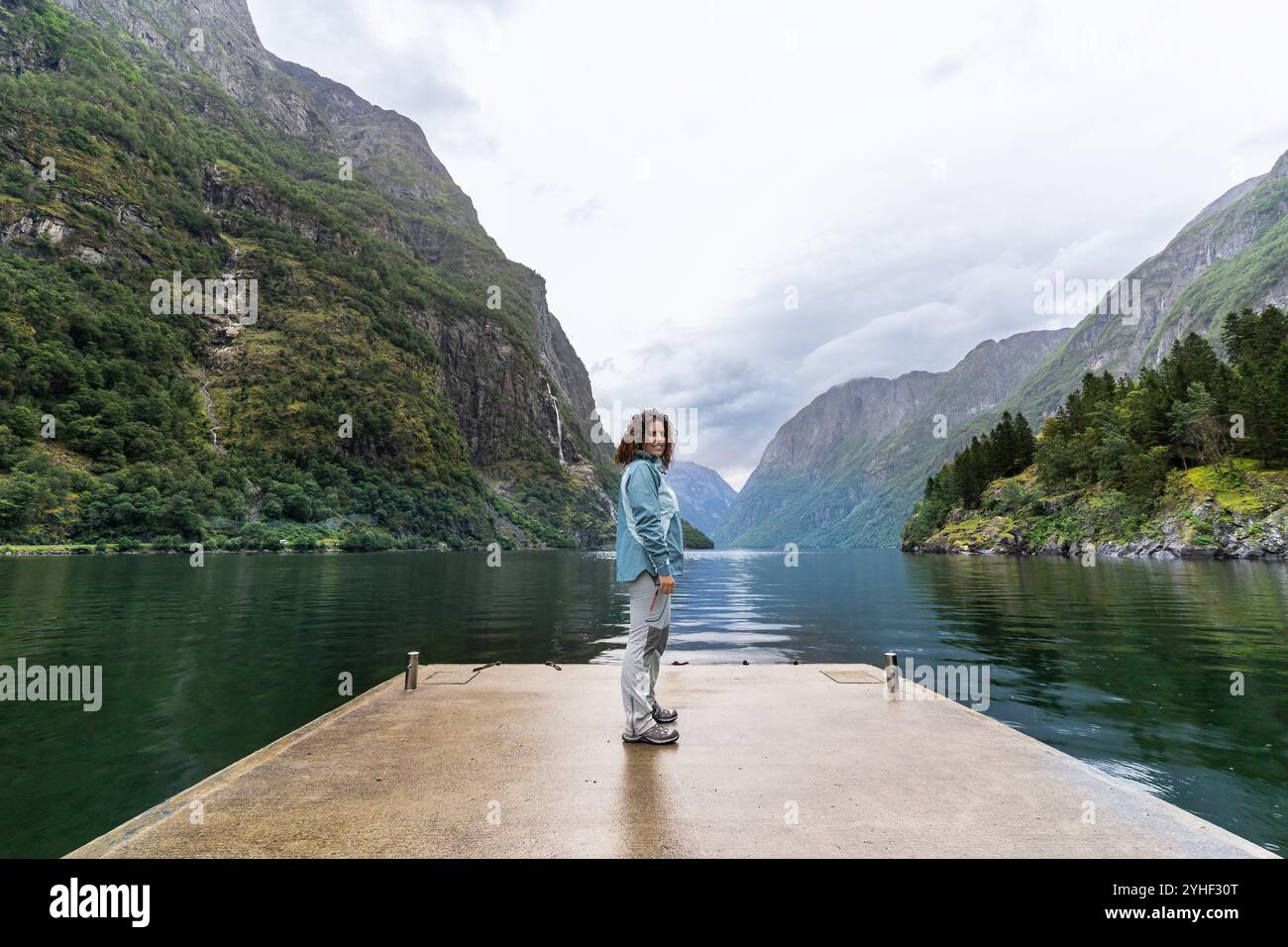 Eine junge Frau, um 30 Jahre alt, steht an einem bewölkten Tag auf einem kleinen Dock und blickt auf die Kamera vor dem Hintergrund der weiten Landschaft des Nærøyfjord. Die Stockfoto