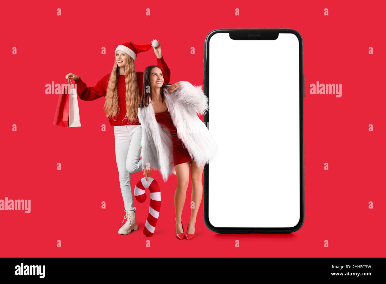 Junge glückliche Frauen mit Einkaufstaschen und großem Smartphone auf rotem Hintergrund. Weihnachtsfeier Stockfoto