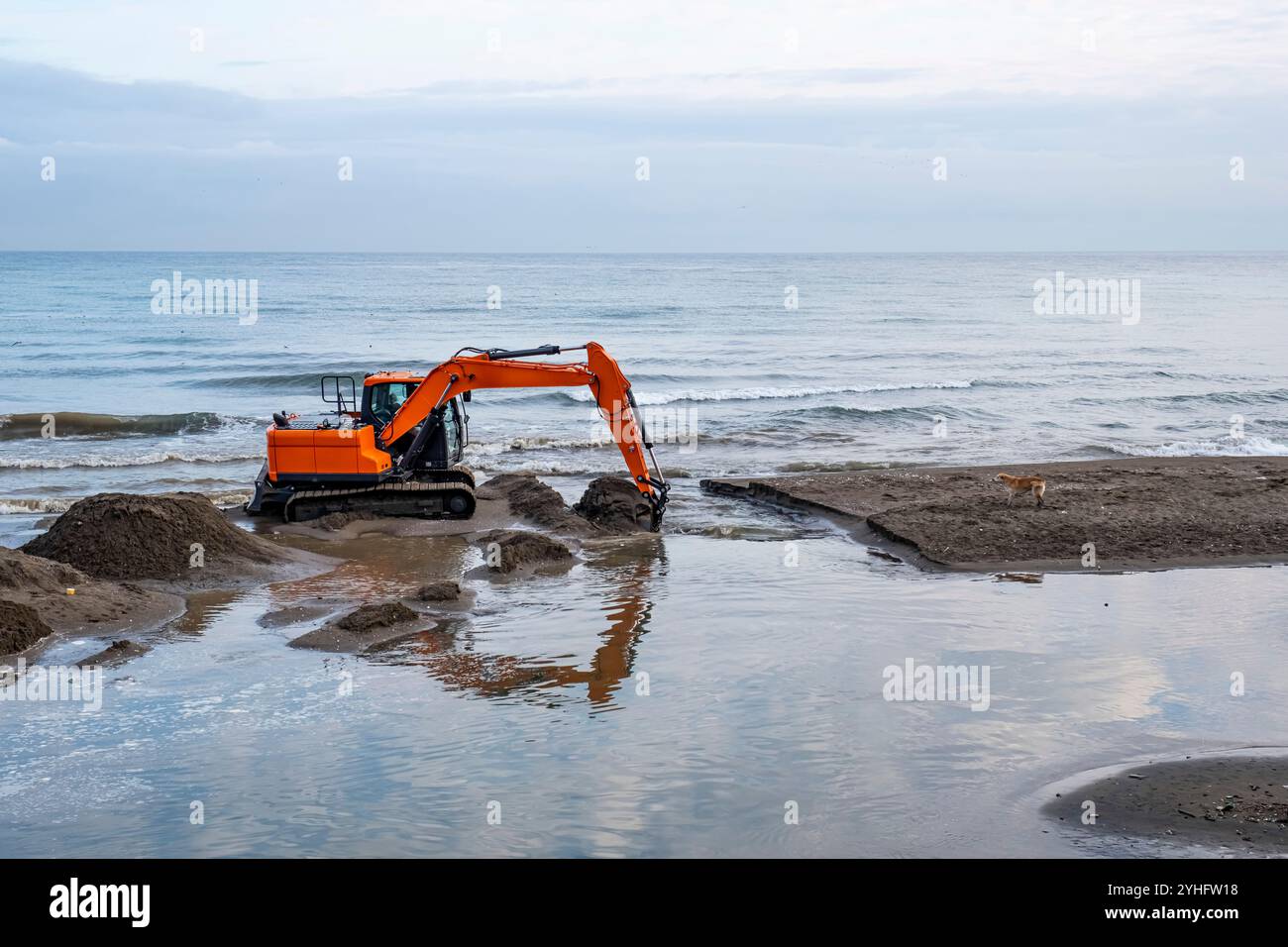 Ein großer orangefarbener Bagger befindet sich am Strand und gräbt sich in den Sand. Der Strand ist nass und das Wasser trüb Stockfoto