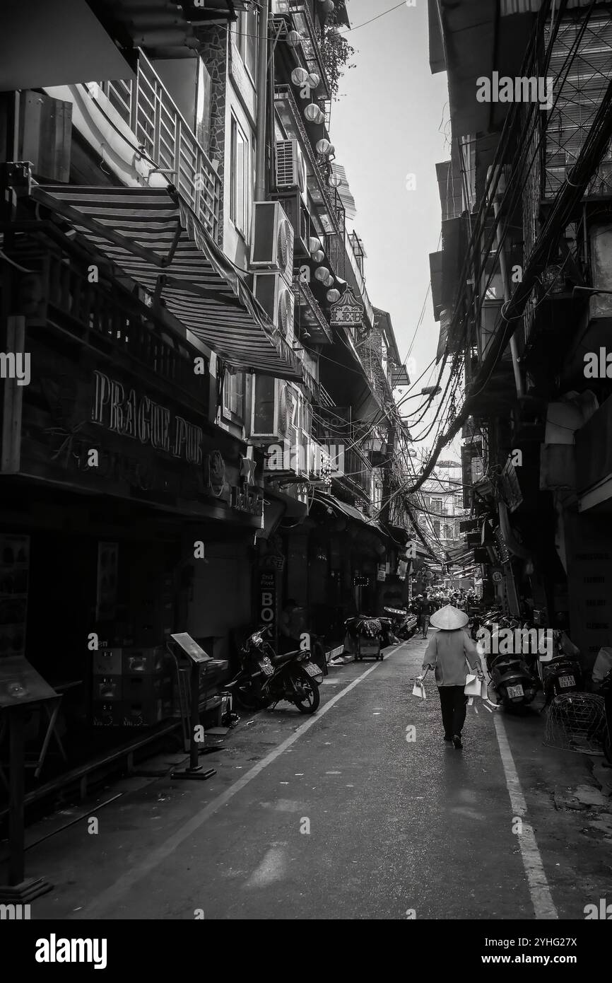 Eine Frau mit einem traditionellen vietnamesischen konischen Hut geht in Hanoi eine enge Straße entlang, vorbei an dicht gepackten Gebäuden und Motorrädern. Stockfoto