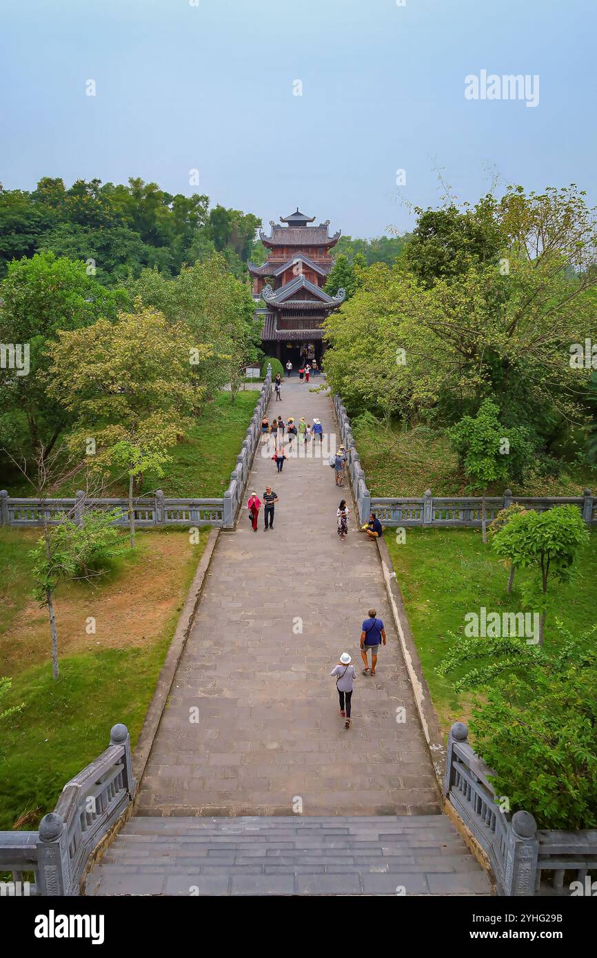 Besucher gehen auf einem Weg zum Haupteingang des Bai Dinh Tempels, flankiert von üppigem Grün und traditioneller vietnamesischer Architektur. Stockfoto