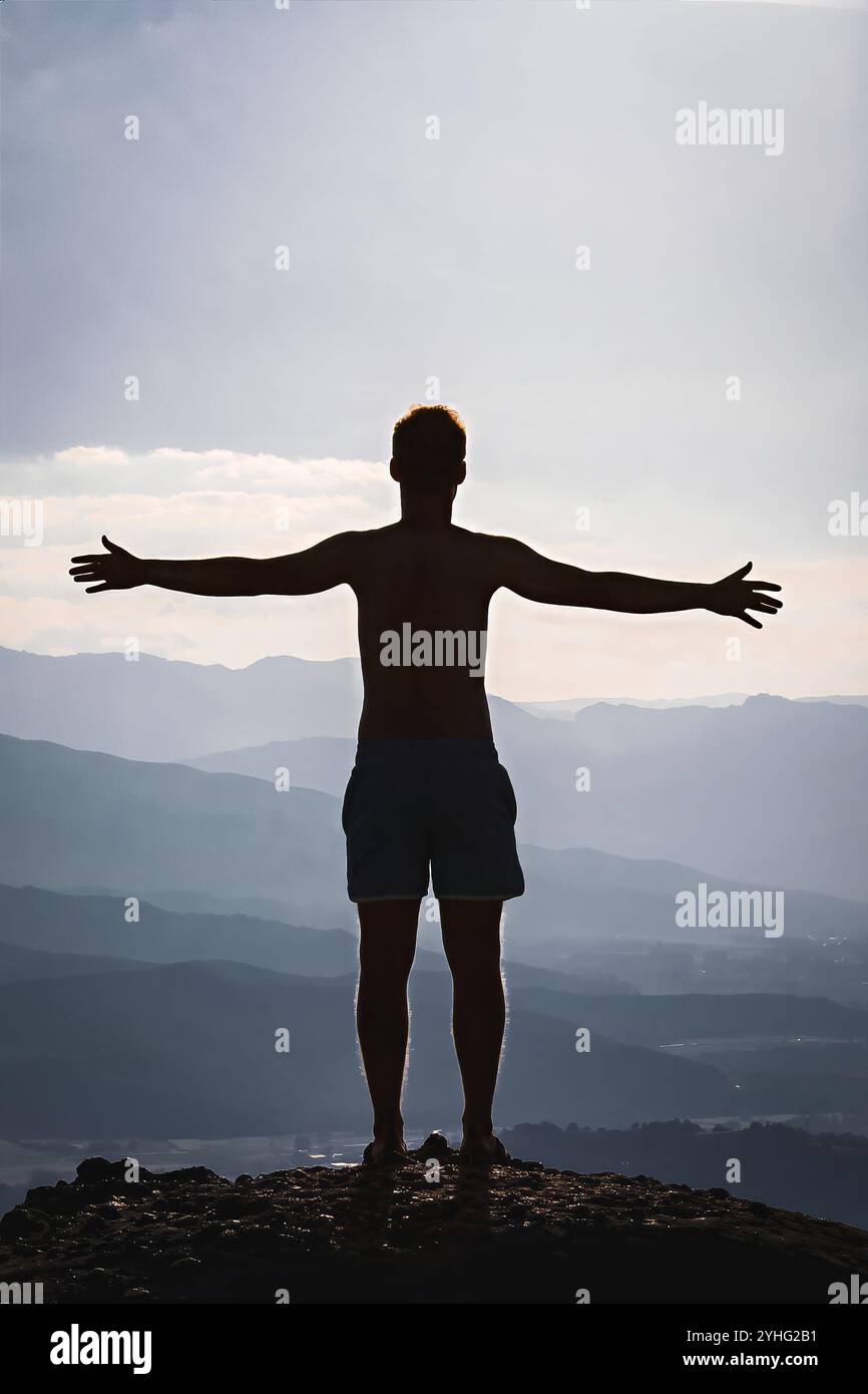 Silhouette eines Mannes mit offenen Armen, der auf einem Berggipfel steht und die Weite der Natur umhüllt, mit Schichten von Bergen, die sich bis zum Horizont erstrecken. Stockfoto