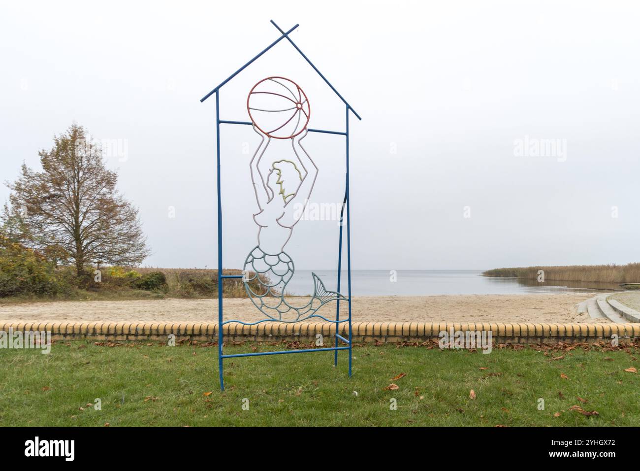 Eine dekorative Metallskulptur einer Meerjungfrau, die einen Ball hält, vor der ruhigen Kulisse von Stettiner Haff an einem bewölkten Novembertag. Stockfoto