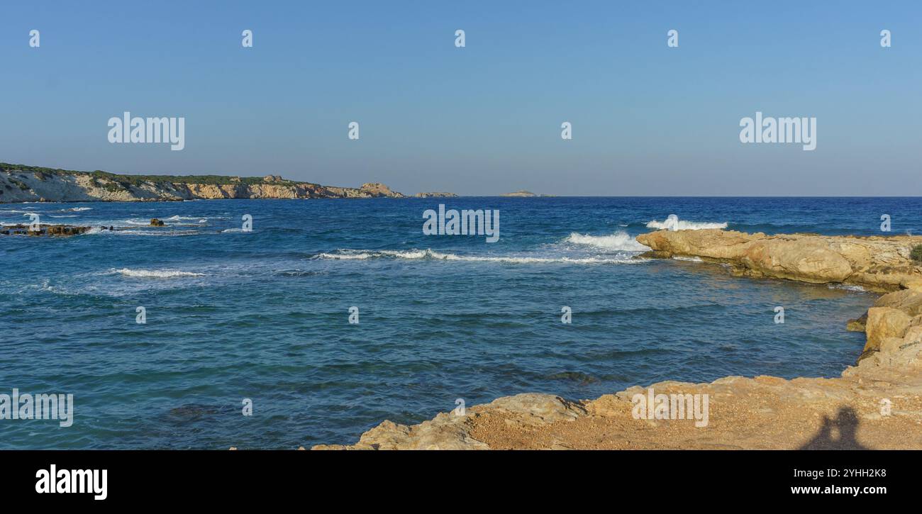 Die ruhige Küstenlandschaft bietet strahlend blaues Wasser und sanfte Wellen vor sonnendurchfluteten Felsen in der Abenddämmerung. Stockfoto