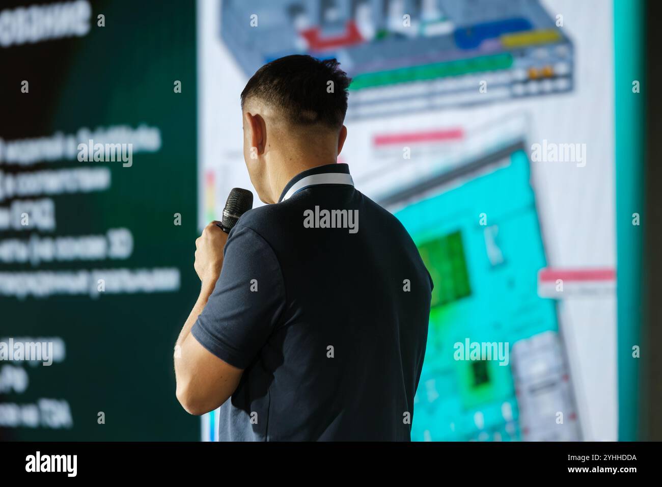 Geschäftsmann mit Mikrofon, der Daten auf einem großen Bildschirm präsentiert. Professionelles Umfeld mit Schwerpunkt auf digitaler Informationspräsentation und Kommunikationsfähigkeiten. Stockfoto