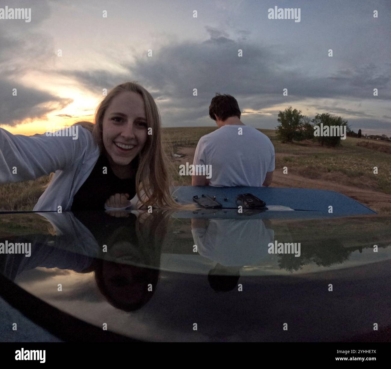 Ein Paar genießt einen ruhigen Sonnenuntergang, während es auf einer Autohaube in einer ländlichen Landschaft sitzt Stockfoto