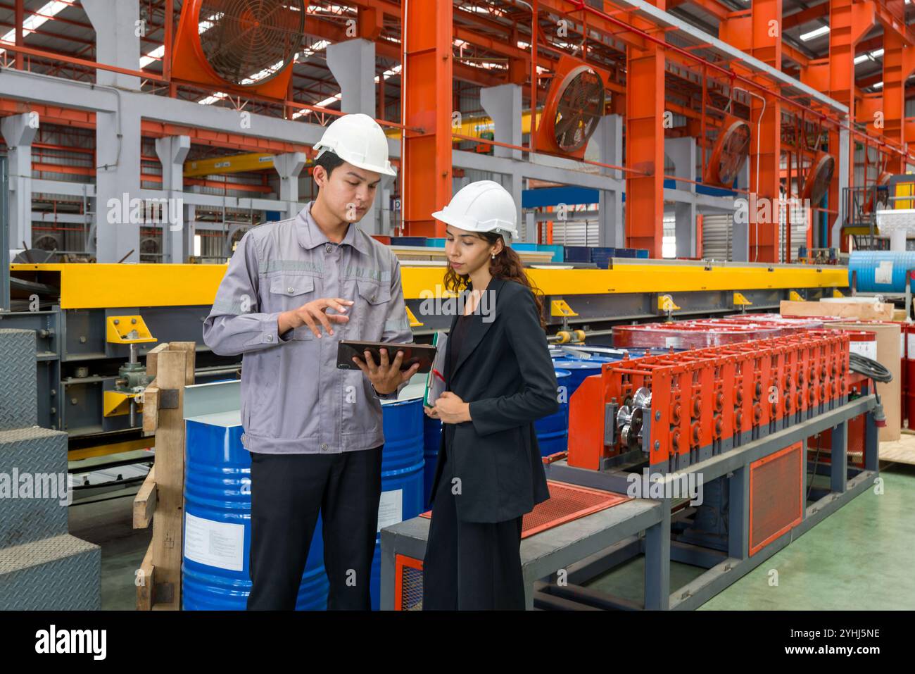 Zwei Arbeiter mit Schutzhelm und schwarzem Anzug stehen in einer Fabrik, halten einen Tablet-Computer in der Hand, um Notizen zu machen, den Zeitplan zu überprüfen, Digitalanzeige zu überprüfen Stockfoto