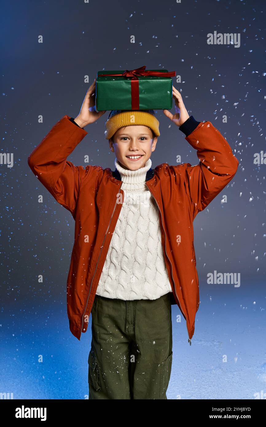 Ein Junge in einem gemütlichen Pullover und einer orangefarbenen Jacke hält glücklich ein Geschenk über dem Kopf im Schnee. Stockfoto