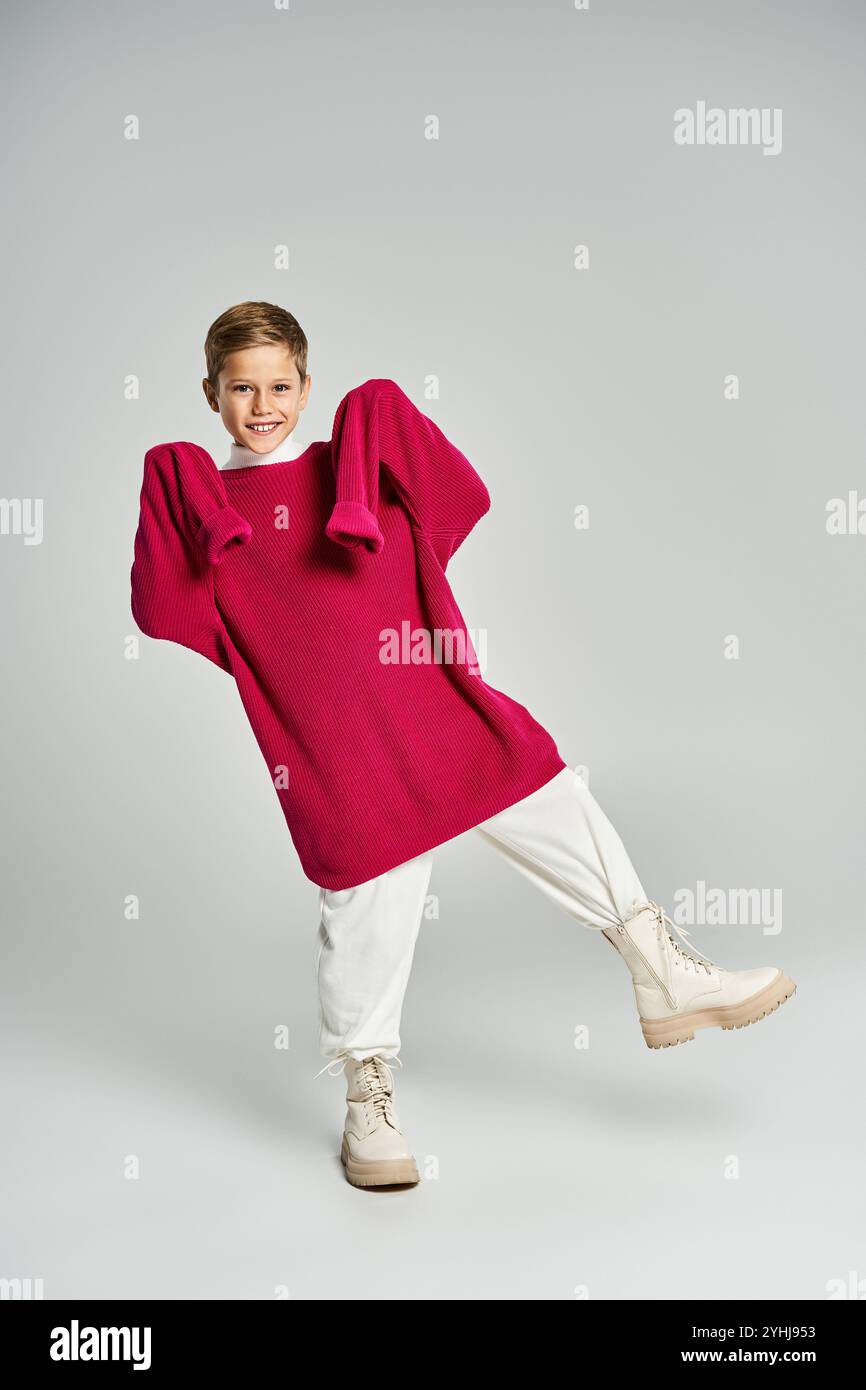 Ein fröhlicher Junge modelliert einen lebendigen, übergroßen Pullover, während er im Winter eine lustige Pose zeigt. Stockfoto