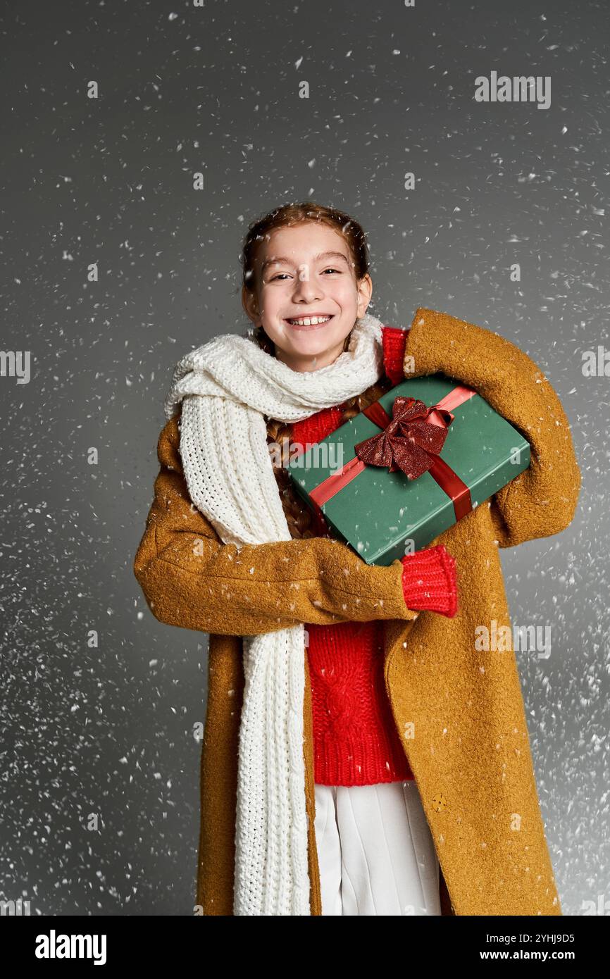 Ein entzücktes Mädchen in warmen Winterkleidung hält ein Geschenk, während Schneeflocken um sie fallen. Stockfoto