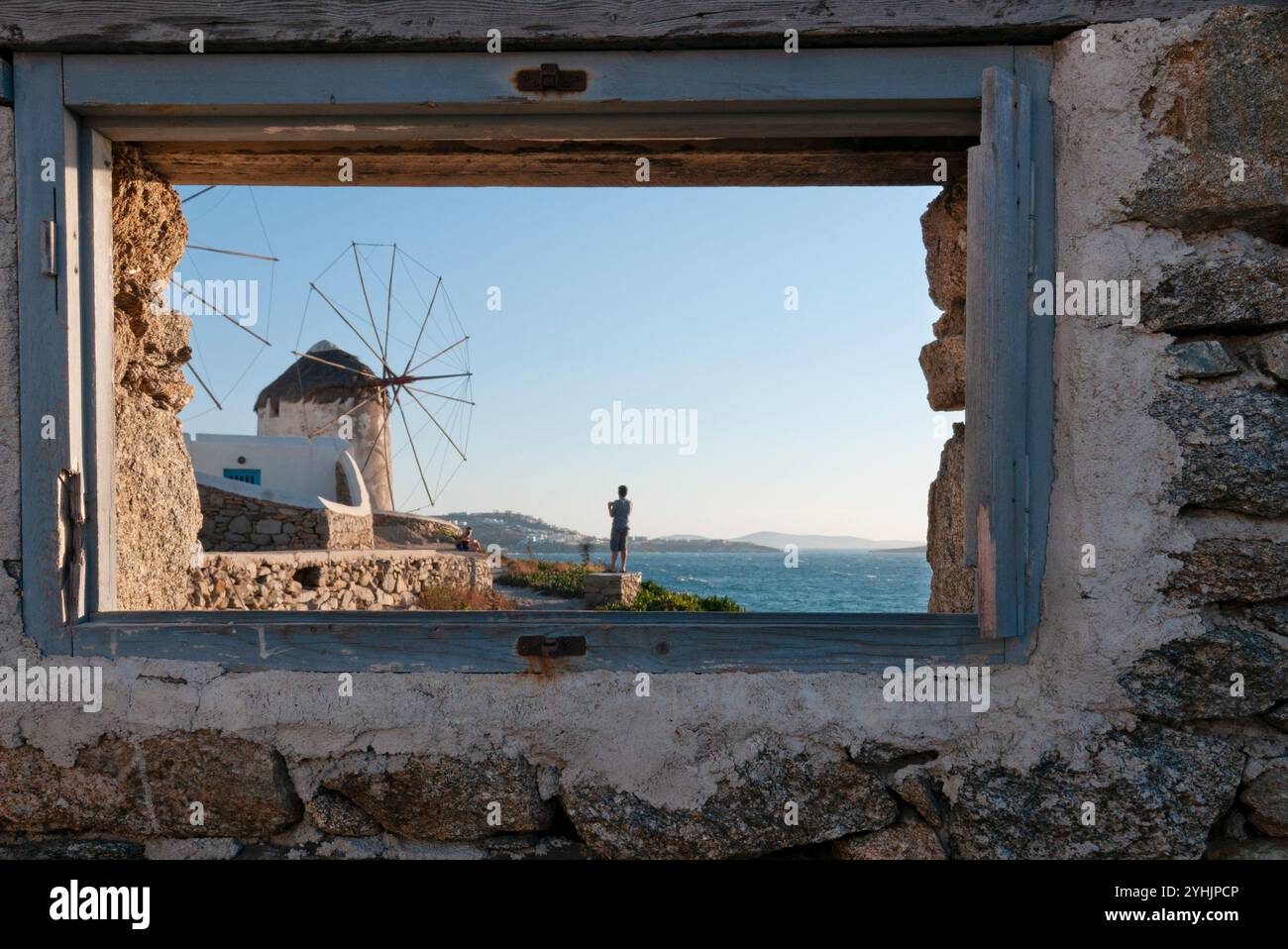 Traditionelle griechische Windmühle am Meer auf Kykonos Insel, eingerahmt von einem rustikalen Fenster in einer Steinmauer, beobachtet ein Tourist die Landschaft bei Sonnenuntergang Stockfoto