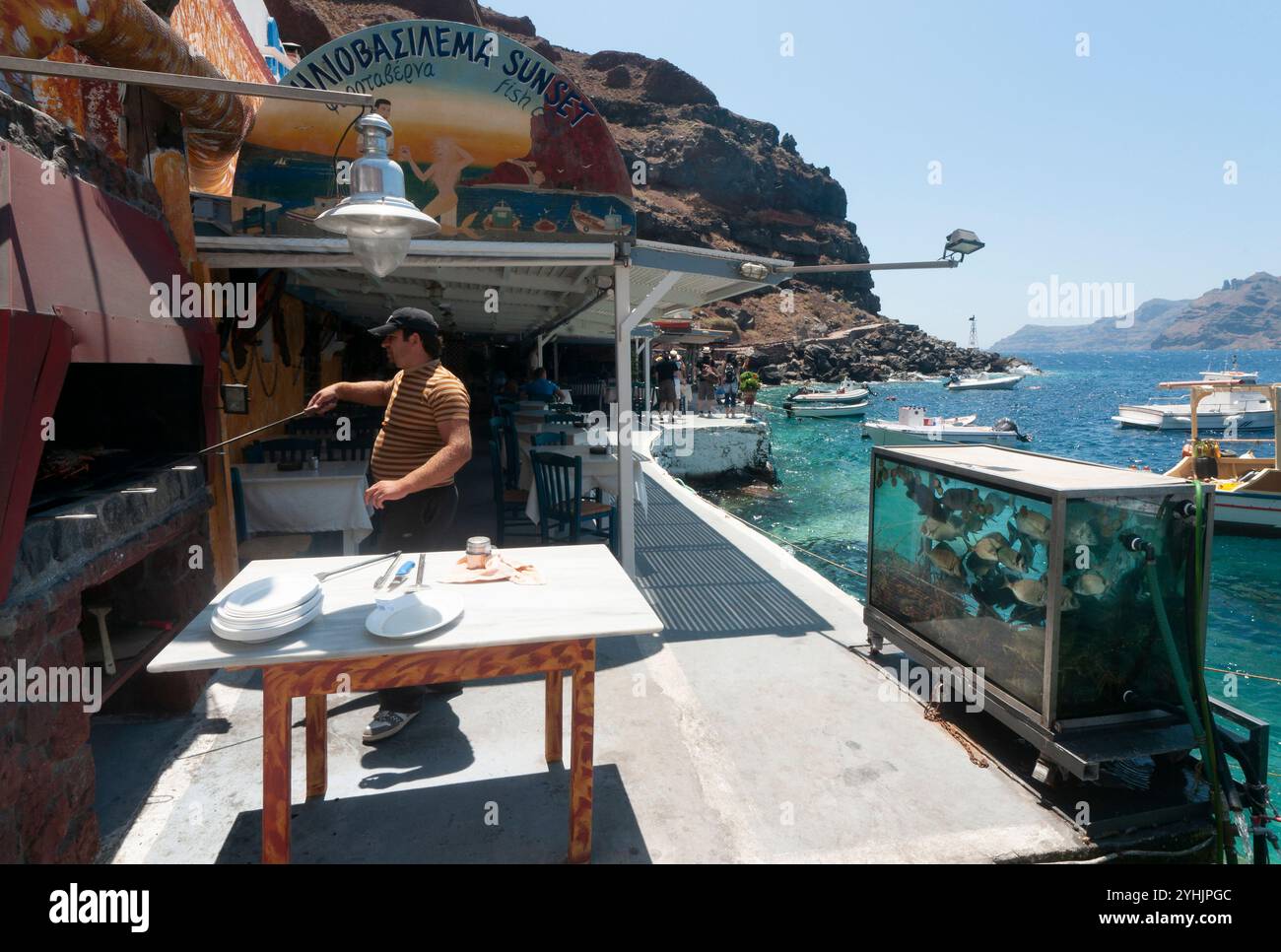 Mann grillt in einem Restaurant am Meer in der Bucht von Ammoudi mit einem „Sonnenuntergang“-Schild in griechischer Sprache, umgeben von Gerichten, Aquarium, Booten auf dem Wasser und bergig Stockfoto