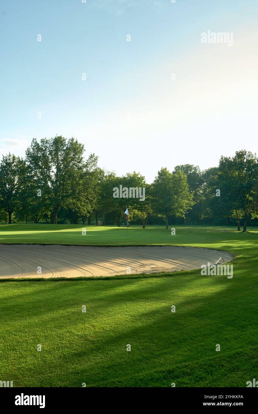 Ruhiger Golfplatz mit Sandbunker und Flagge, umgeben von üppigen Bäumen unter einem klaren blauen Himmel. Perfekt für Konzepte der Entspannung, der Natur und des Outdoo Stockfoto