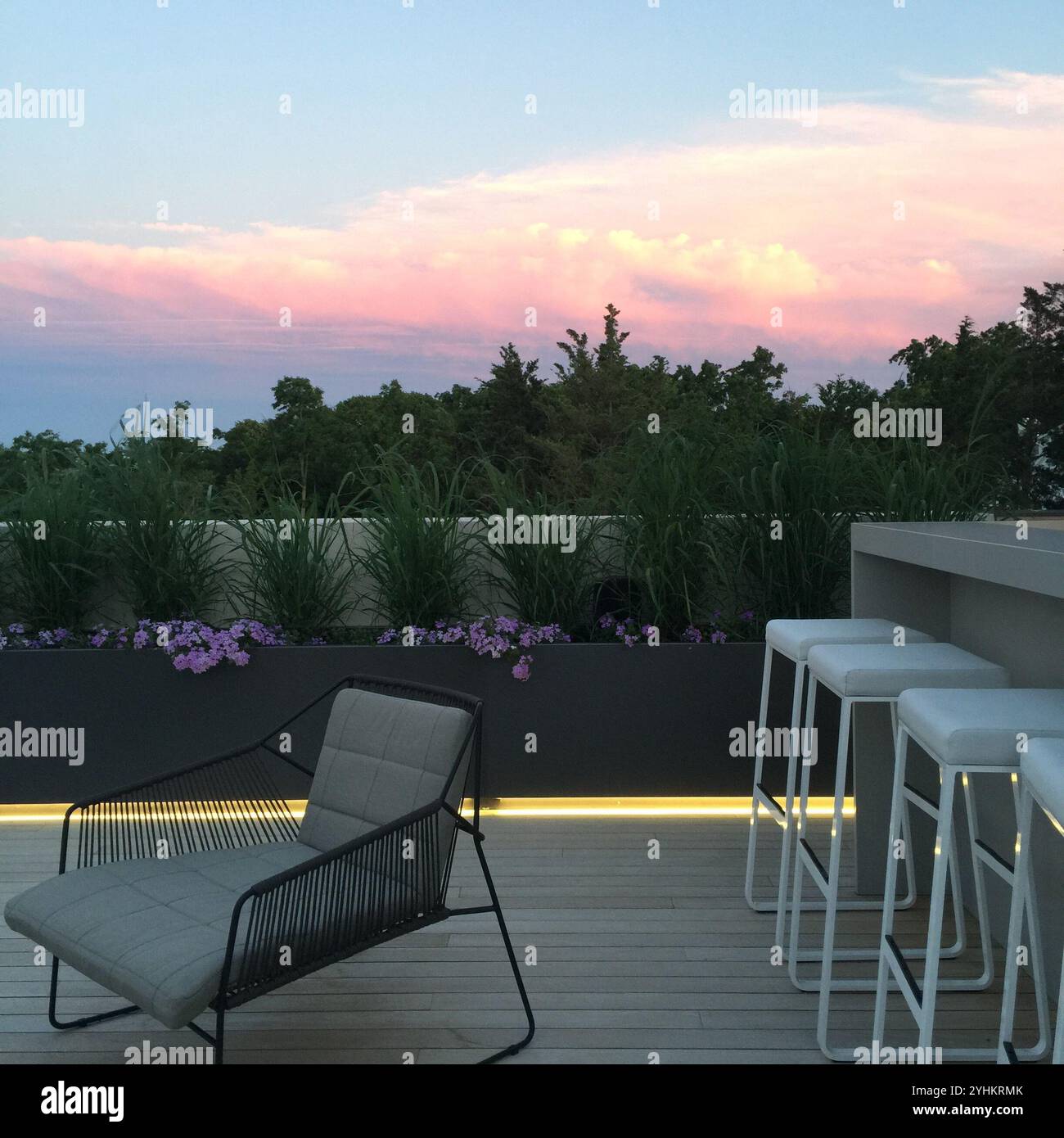 Lounge auf der Dachterrasse bei Sonnenuntergang mit elegantem Sessel und modernen Barhockern. Rosafarbene Wolken leuchten am Abendhimmel, während Grün und violette Blüten entstehen Stockfoto