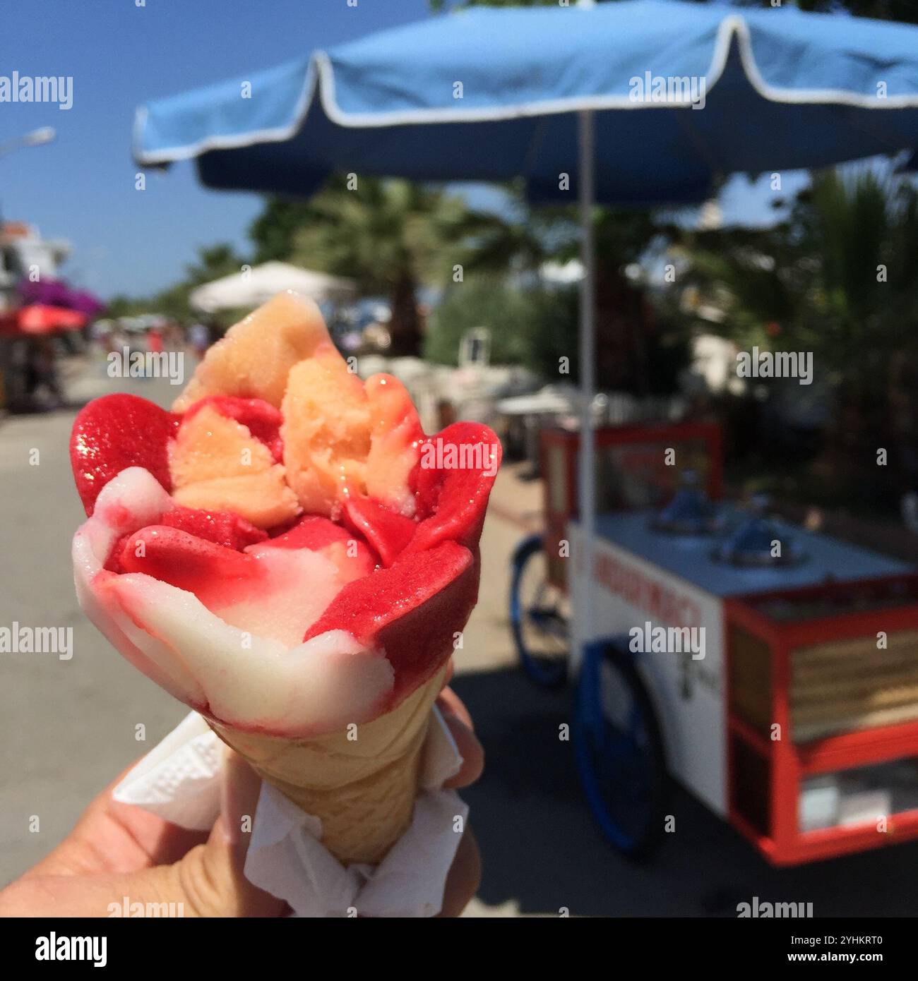Eine erfrischende Eiskegel mit leuchtenden roten und pfirsichfarbenen Aromen, die wie eine Blume geformt sind, wird an einem sonnigen Tag von einem Einkaufswagen gehalten. Stockfoto