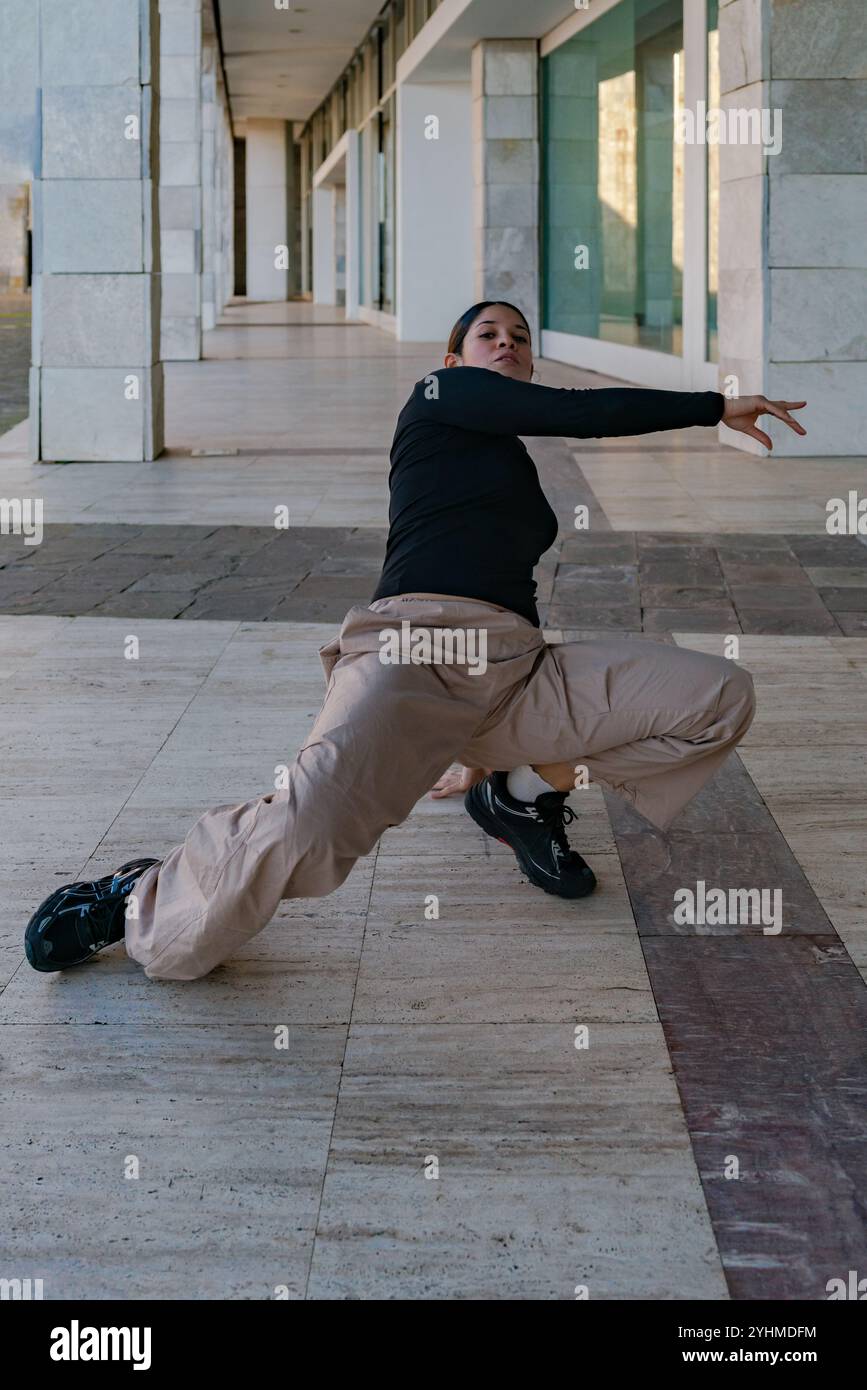 Eine junge Frau posiert beim Breakdance auf einem Bürgersteig. Sie trägt ein schwarzes T-Shirt und eine braune Hose Stockfoto