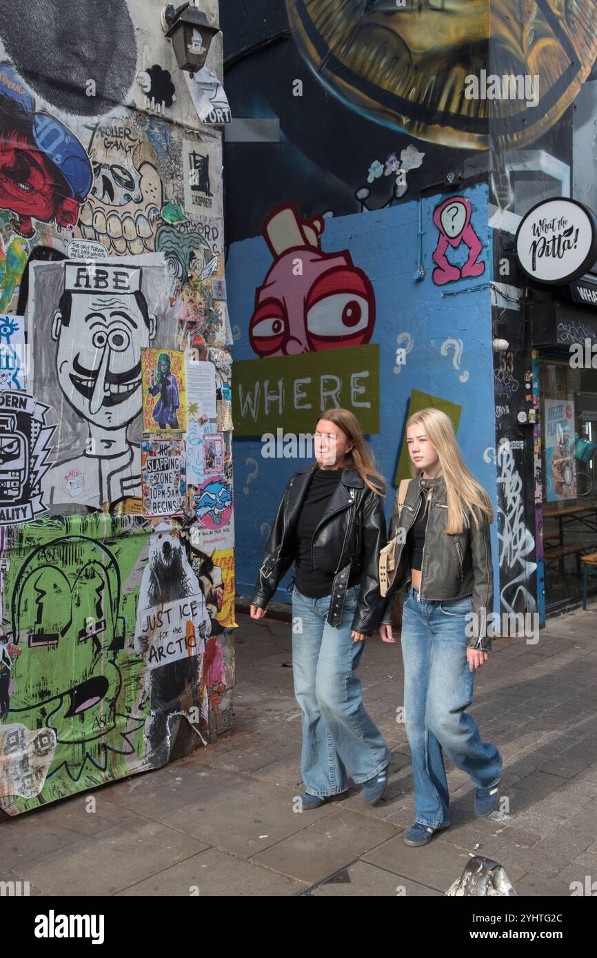 Street Fashion Style London 2020s UK. Brick Lane East End London Mutter und Erwachsene Tochter, die ähnliche Kleidung trägt. Schwarze Lederjacken, verblasst, gebraucht, Glockenfackeln. Blaue Jeans und blaue Sneakers. Beide haben lange blonde Haare. Sie gehen an einer Wand vorbei, die mit Graffiti-Wandkunst bedeckt ist. Brick Lane, Tower Hamlets, East End London England 2024 HOMER SYKES Stockfoto