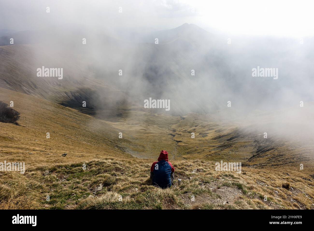 Ein einsamer Wanderer in roter Jacke, der auf einem grasbewachsenen Berghang sitzt, blickt über das nebelige Tal mit Nebel über die Landschaft vom Moldoveanu Gipfel, Rumänien Stockfoto