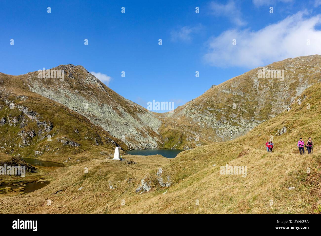 Wanderer, die auf einem grasbewachsenen Bergpfad neben einem abgelegenen Bergsee wandern, umgeben von felsigen Gipfeln unter einem klaren blauen Himmel Stockfoto