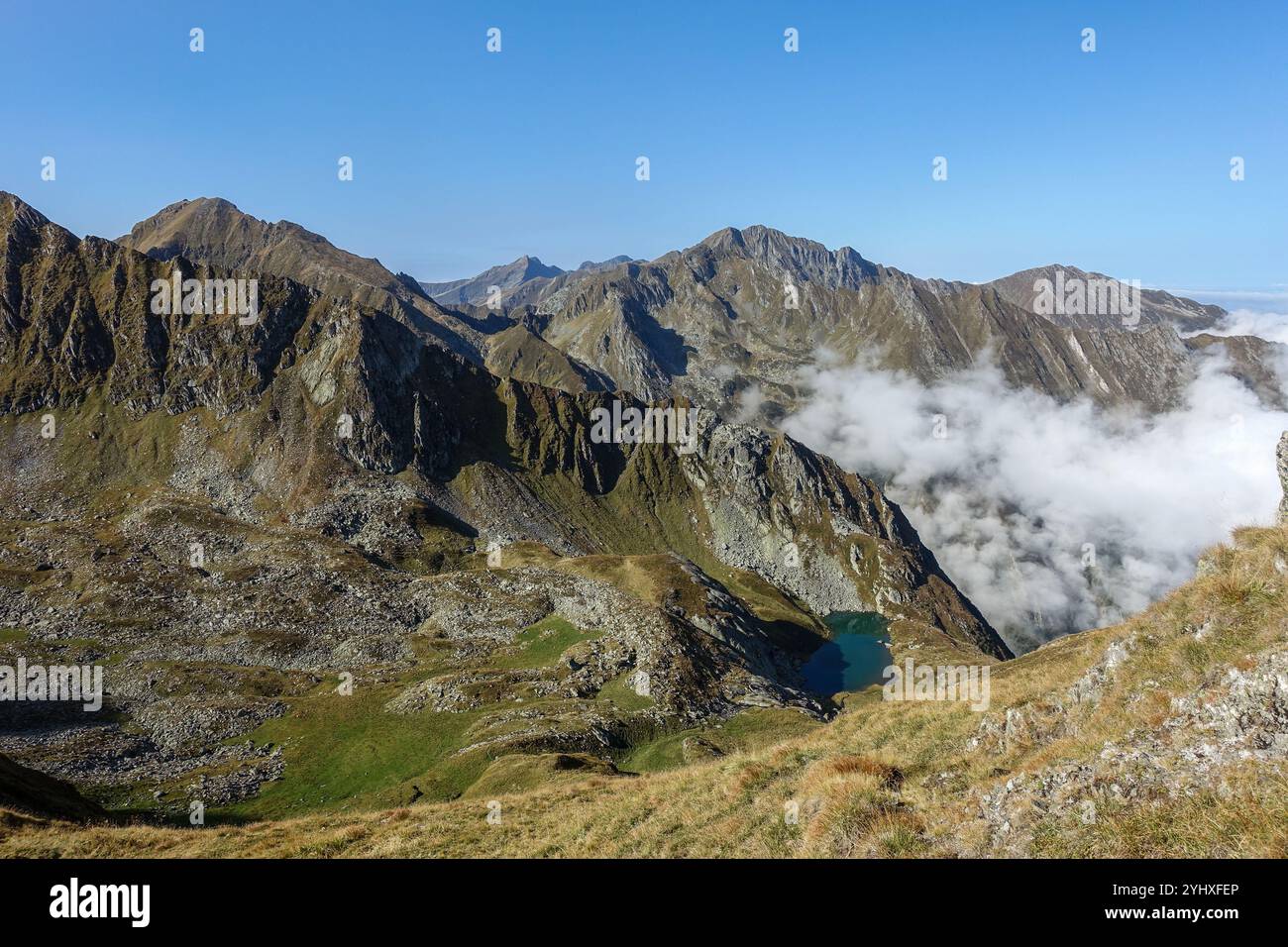 Felsiges Bergmassiv mit steilen Bergrücken und einem kleinen alpinen See unter einem klaren blauen Himmel und treibenden Wolken, Moldoveanu Trail, Rumänien Stockfoto