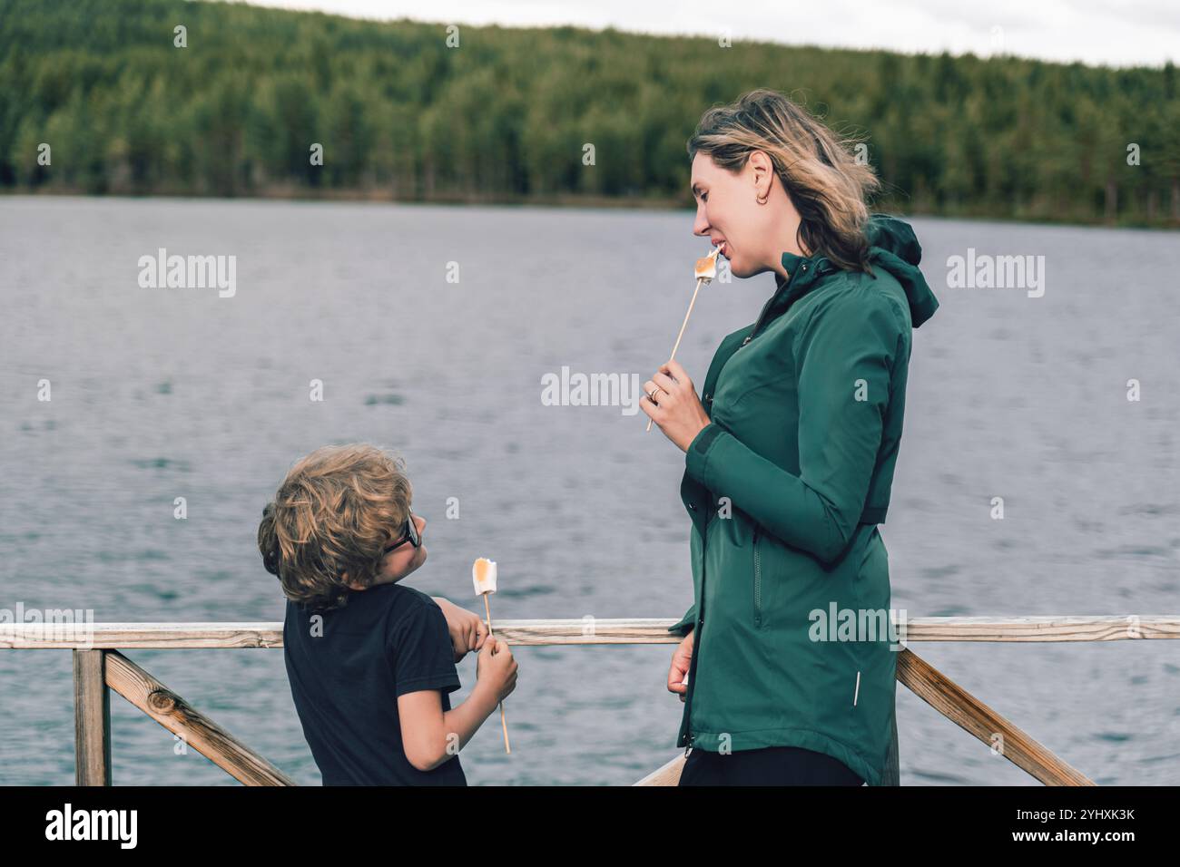 Mutter und Sohn sehen sich fröhlich an, während sie geröstete Marshmallows draußen an einem ruhigen See essen, was einen herzerwärmenden Moment in der Natur schafft Stockfoto