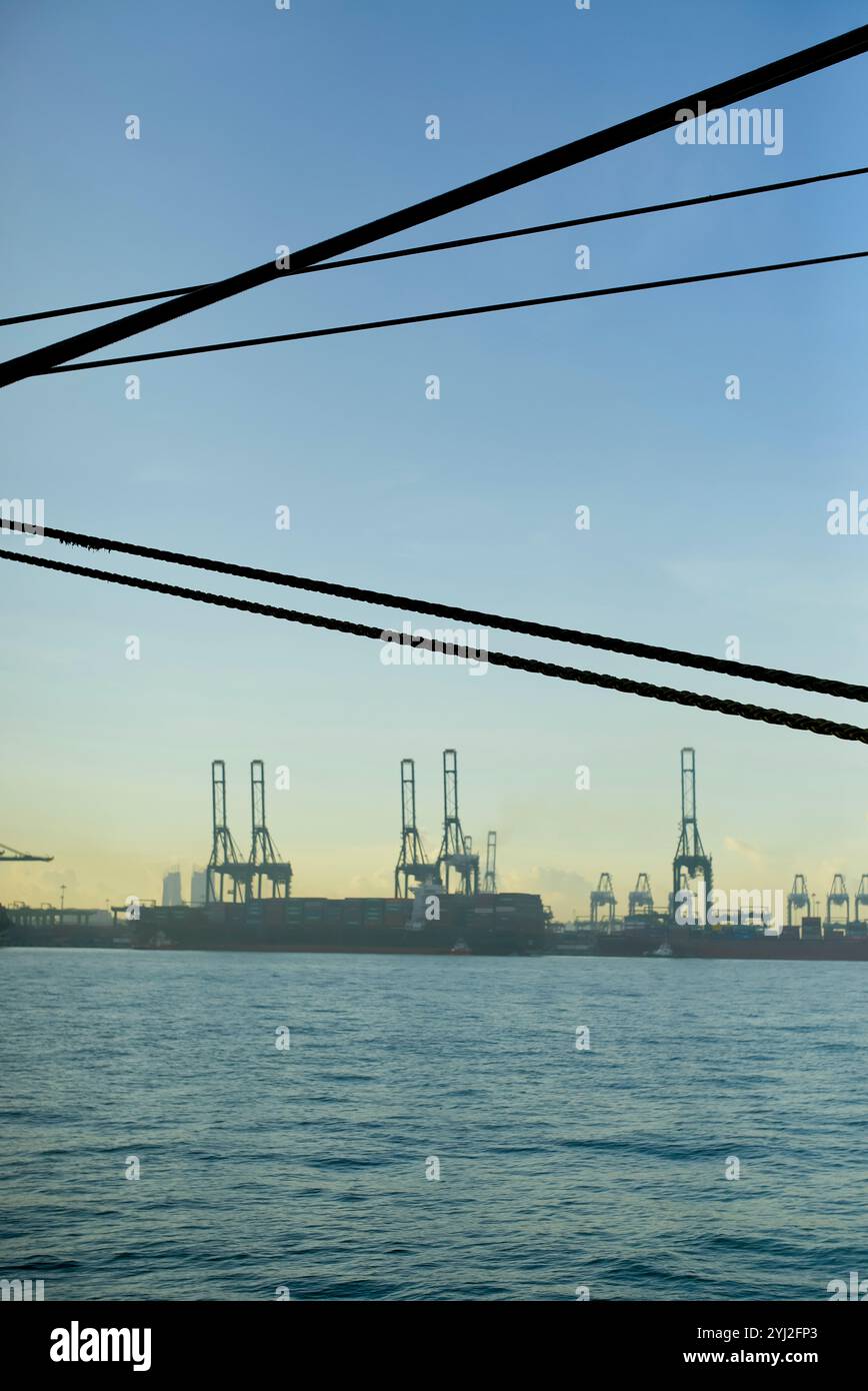 Silhouettenkrane an einem Schiffshafen vor klarem Himmel bei Sonnenauf- oder -Untergang, mit sichtbaren Stromleitungen im Vordergrund, Singapur Stockfoto