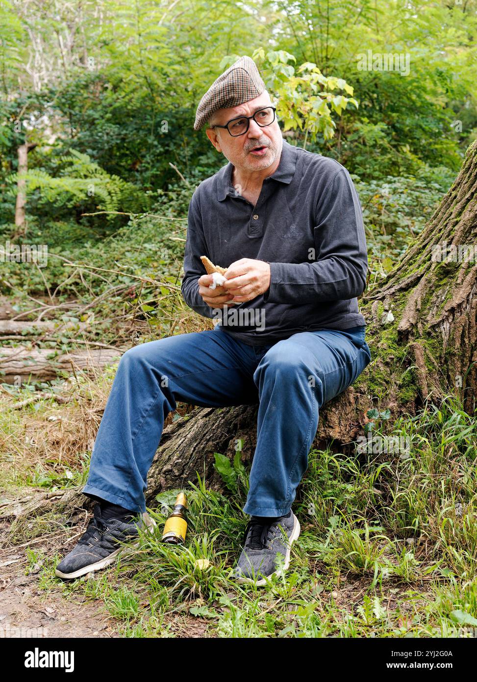 Mann in Brille und Tweedmütze, der auf einem umgestürzten Baumstamm sitzt und einen Apfel in einem Waldgebiet schält, Italien Stockfoto Mann in Brille und Tweedmütze, der auf einem umgestürzten Baumstamm sitzt und einen Apfel in einem Waldgebiet schält, Italien Stockfoto