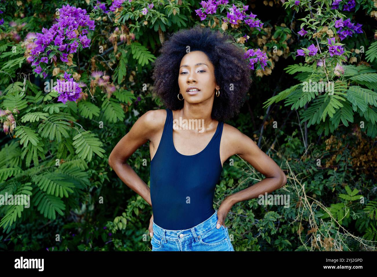 Selbstbewusste Frau mit Afro-Haaren, die vor dem Hintergrund leuchtender lila Blumen und üppig grünem Laub steht. Stockfoto