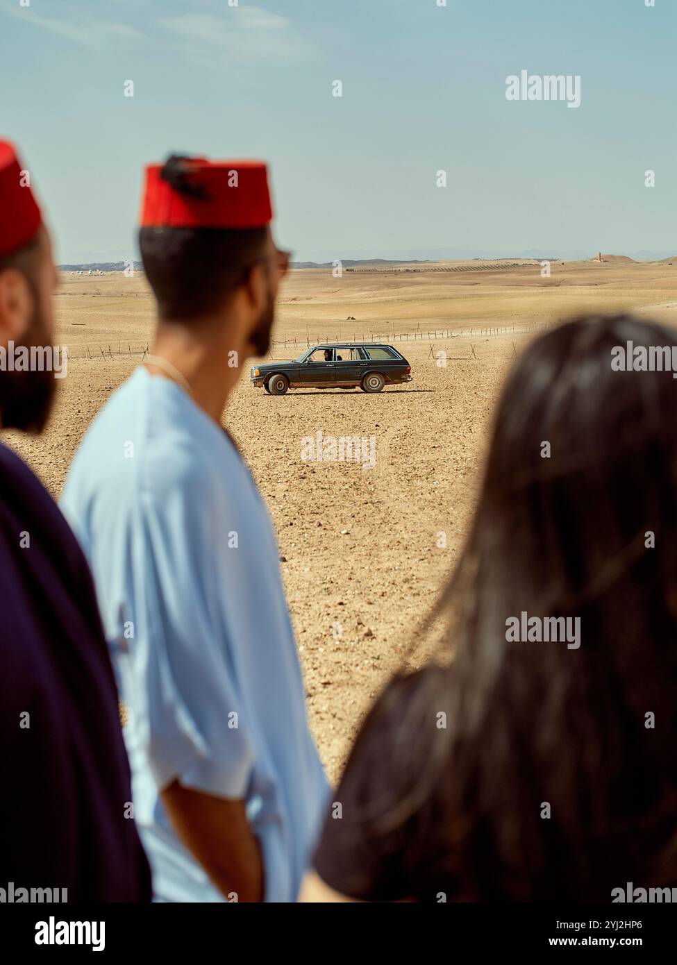 Drei Personen in traditioneller Kleidung beobachten ein Auto in einer Wüstenlandschaft in Marokko Stockfoto