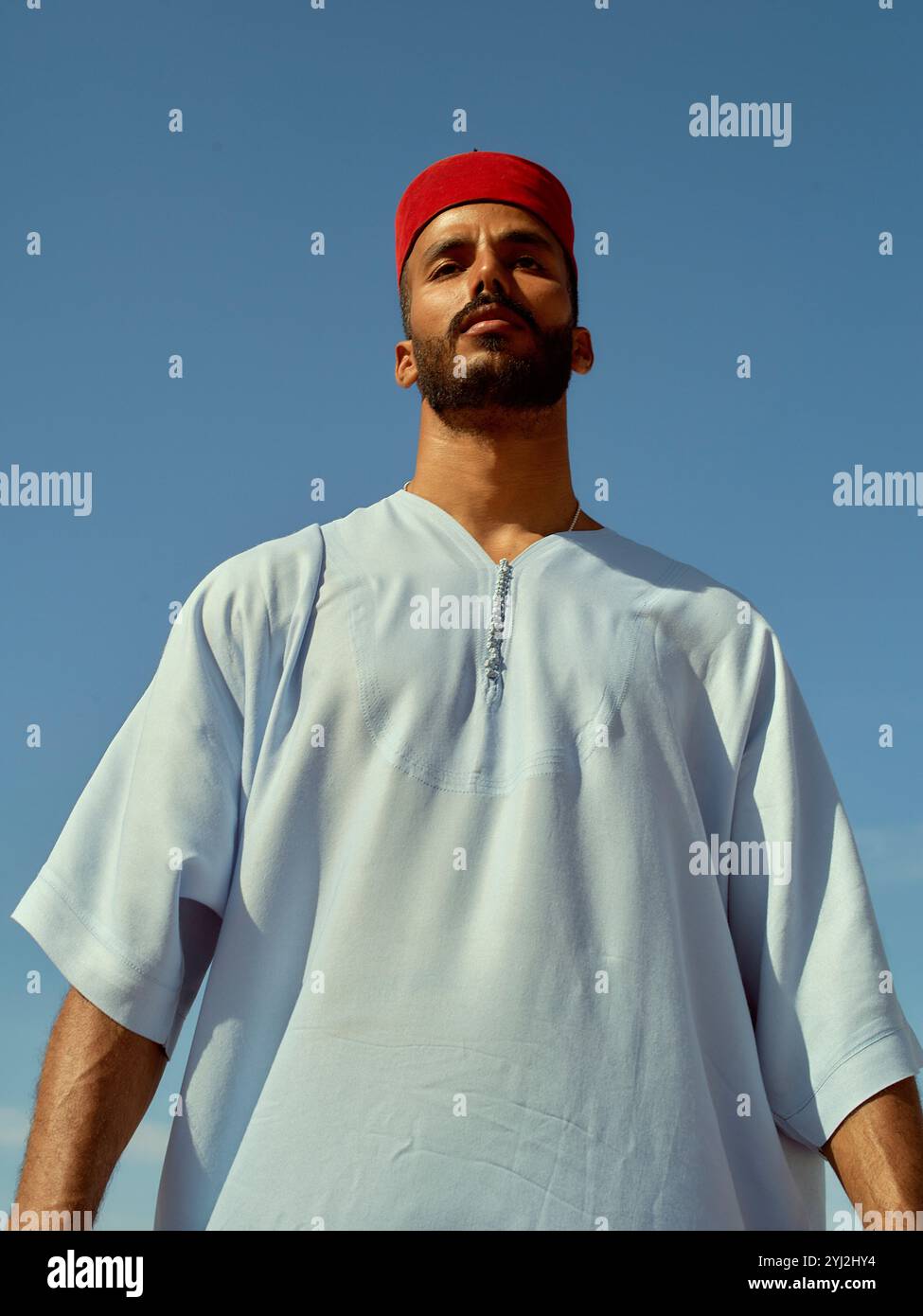 Selbstbewusster Mann mit Bart in traditionellem weißem Gewand und rotem Stirnband vor klarem blauem Himmel, Marokko Stockfoto