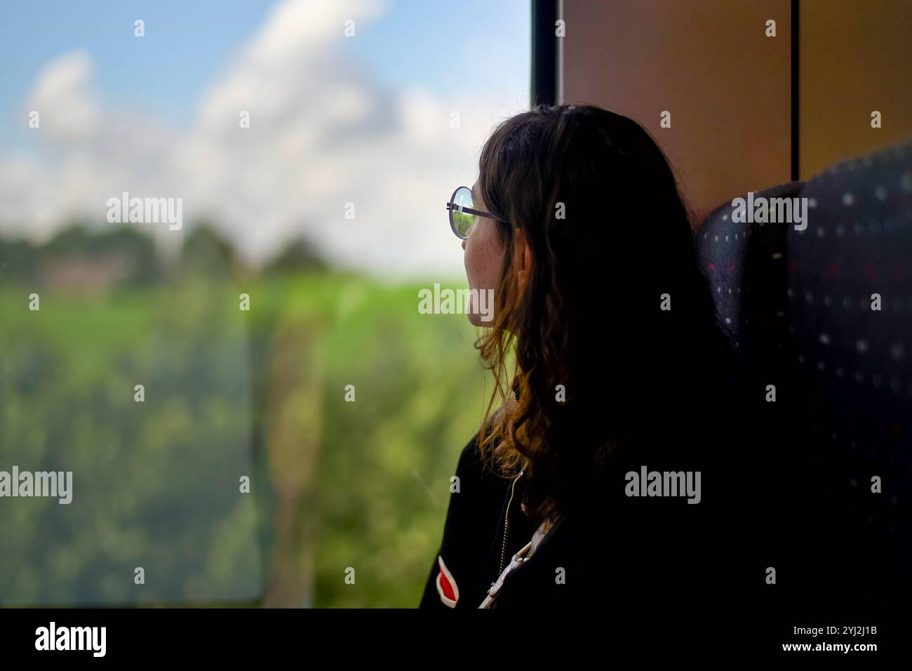 Eine junge Frau mit Brille schaut aus dem Fenster eines Zuges und reflektiert die grüne Landschaft, die vorbeizieht. Stockfoto