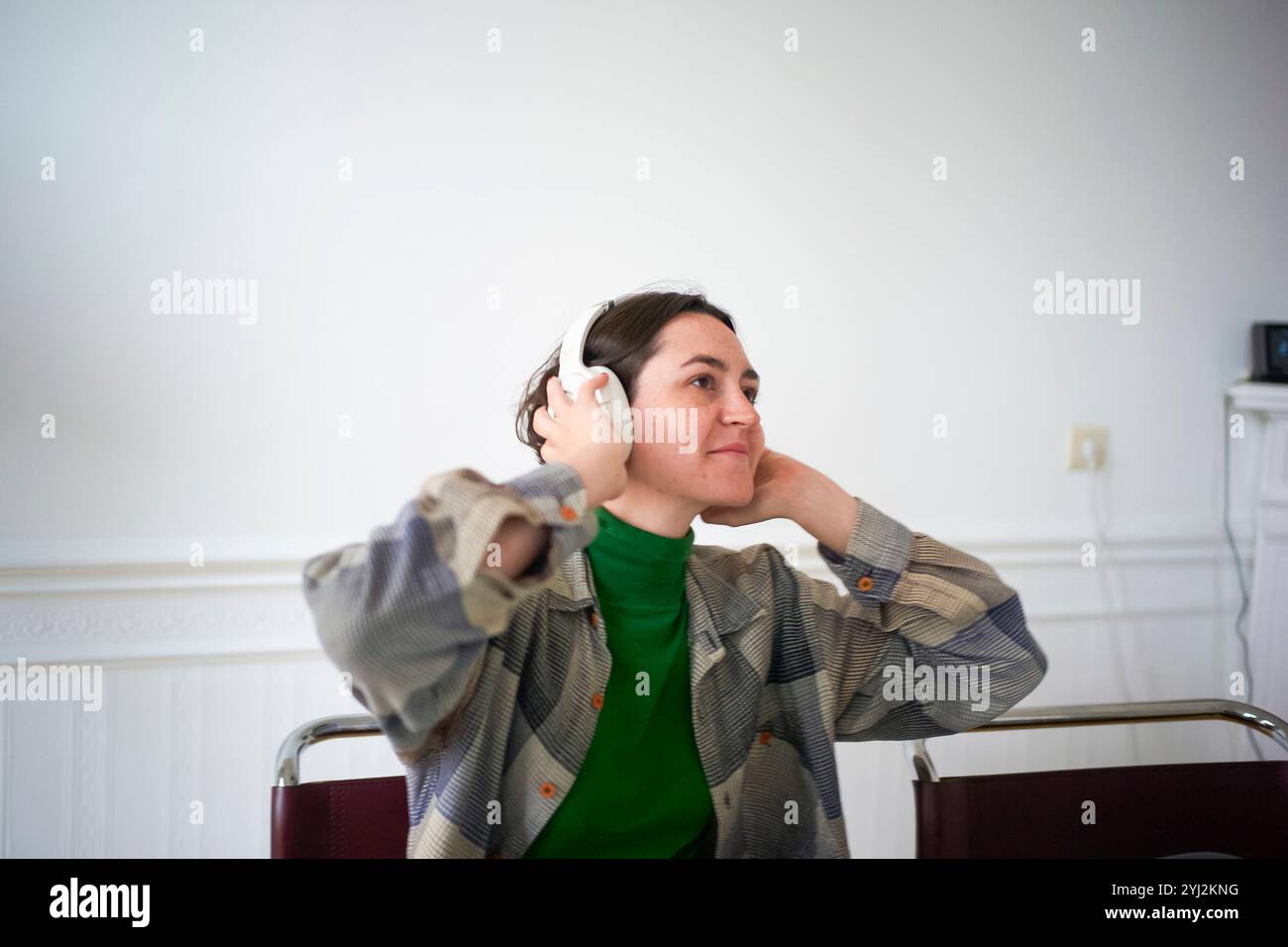 Frau in grünem Oberteil und karierter Jacke, die Musik auf weißen Kopfhörern hört, während sie lächelt und einen Blick auf einen Kollegen wirft Stockfoto