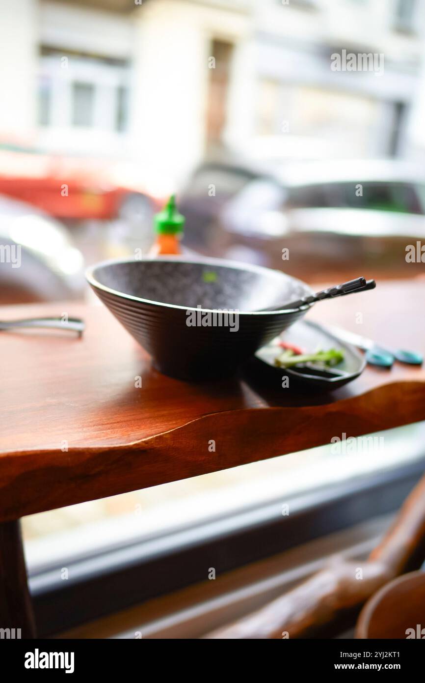 Ein schwarzer leerer Wok und Essstäbchen stehen auf einem Holztisch neben dem Fenster des Restaurants mit Blick auf die Straße. Stockfoto