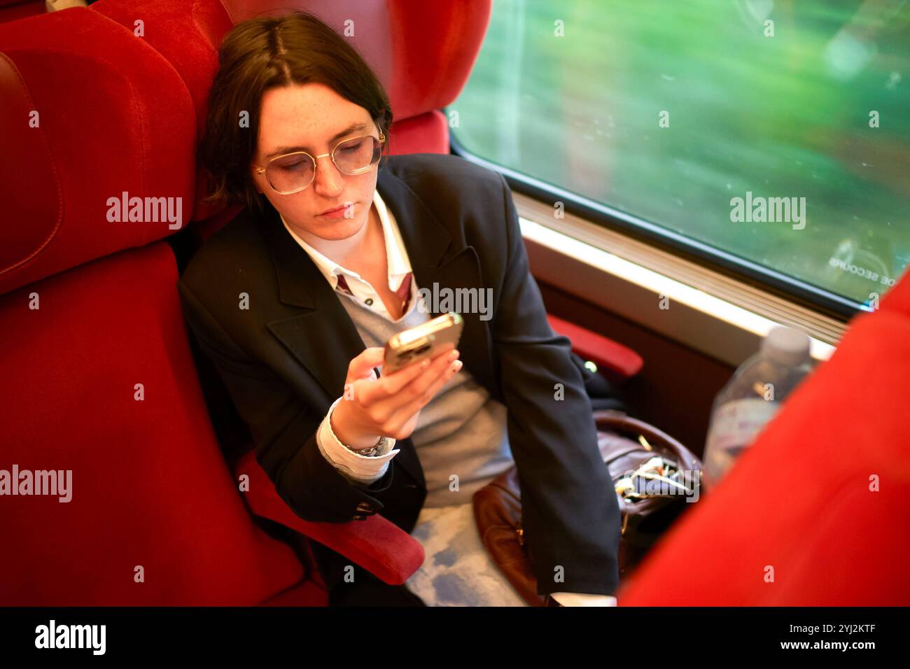 Eine fokussierte junge Frau sitzt auf einem roten Zugsitz, während sie ihr Smartphone während einer Reise durchsucht. Stockfoto